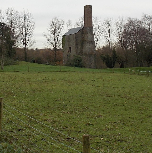 A tall stone engine house with a slate roof and a brick chimney stack rising from it, among bare trees beyond a fenced green field.