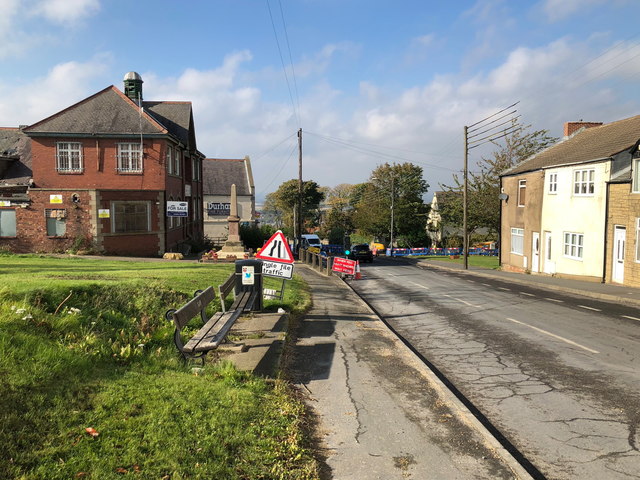 A road running downhill through a former colliery village, with a brick building and a stone memorial on the left, houses on the right and roadworks ahead.
