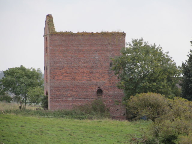 A tall roofless brick engine house standing alone at the edge of a grass field, its gable ragged along the top and a small arched opening low in the front wall.