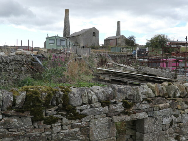 Stone colliery buildings with two tall chimneys seen over a mossy drystone wall, with stacked timber and modern sheds in the yard.
