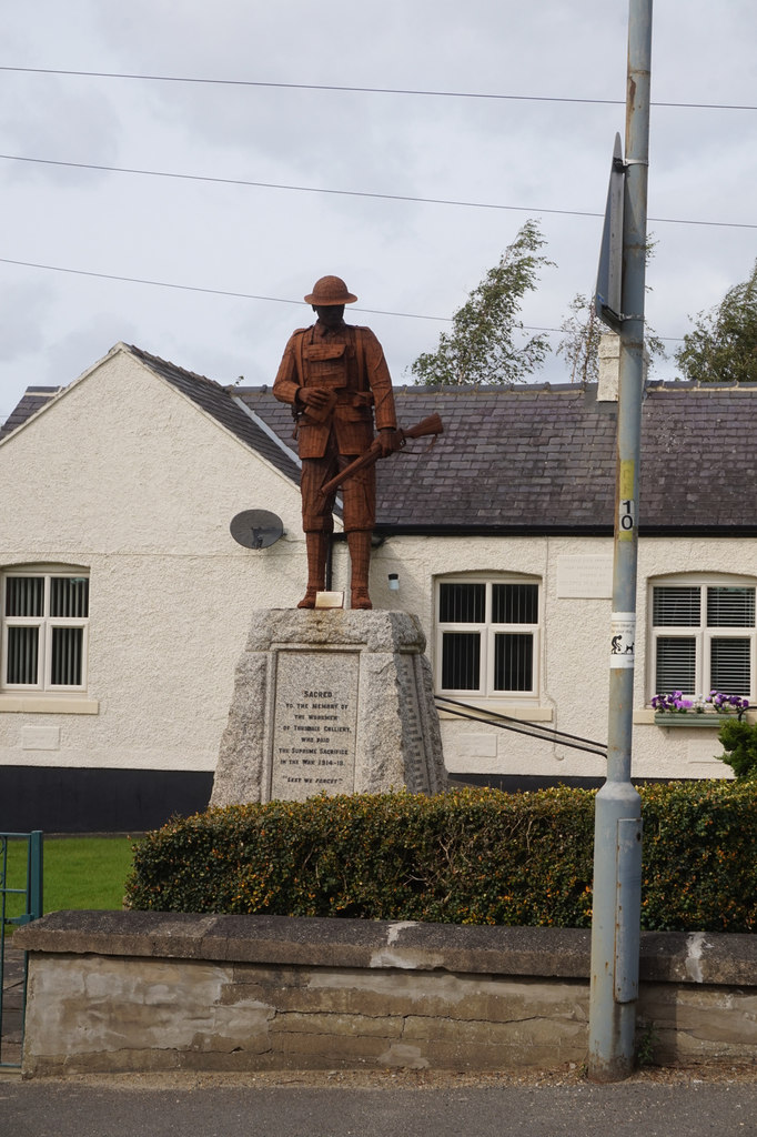 A rust-coloured statue of a soldier with a rifle on an inscribed stone plinth in front of a white cottage.