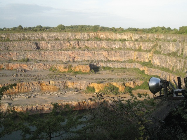 A deep quarry with stepped limestone benches above dark water, seen through trees with a floodlight in the foreground.