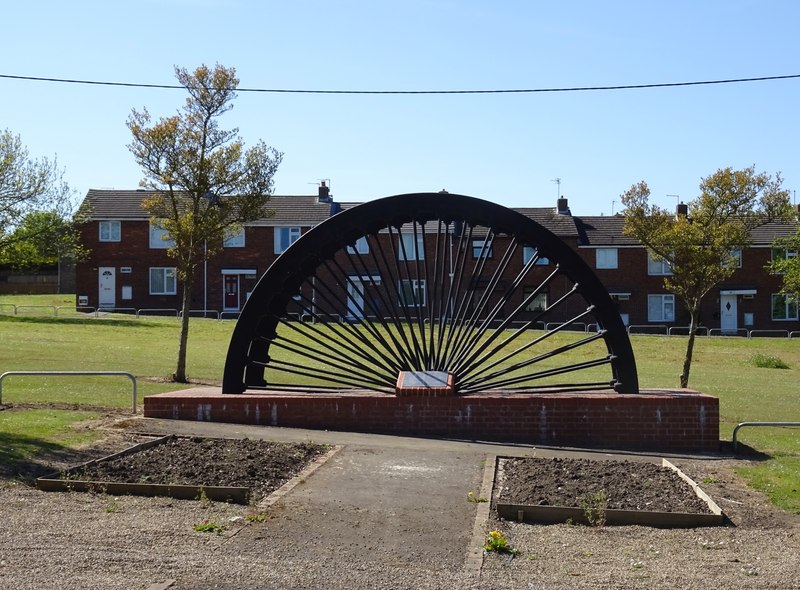 Half of a large black pit winding wheel set upright on a red-brick plinth on a green, with a row of red-brick houses behind, 2025