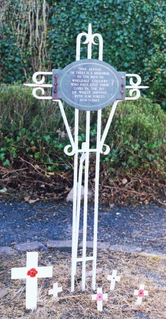 A white-painted wrought-iron memorial cross with an oval plaque, made by the colliery's own blacksmith William Wood, commemorating the men of Wheldale Colliery killed in the pit and those who died serving in the Second World War.