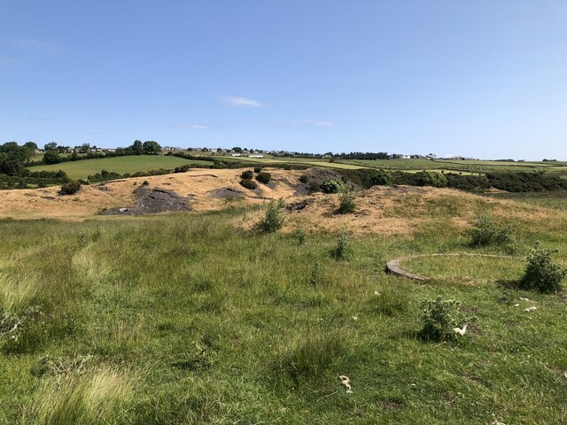 Rough pasture with low grassed mounds, a patch of bare grey shale and a circular concrete ring under a clear blue sky.