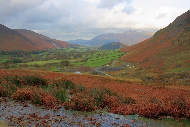 Grey spoil heaps from the lead mine spilling down a grassy fellside above a walled valley, with the bracken-covered flank of the fell behind and the long ridge of Skiddaw on the skyline.