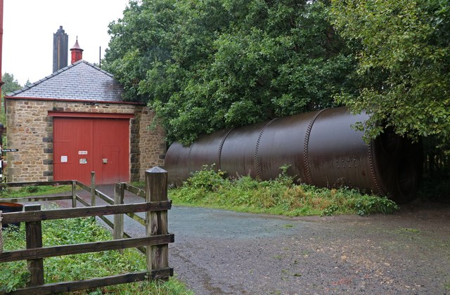A small stone boiler house with red double doors beside a long rusty cylindrical boiler lying among trees.
