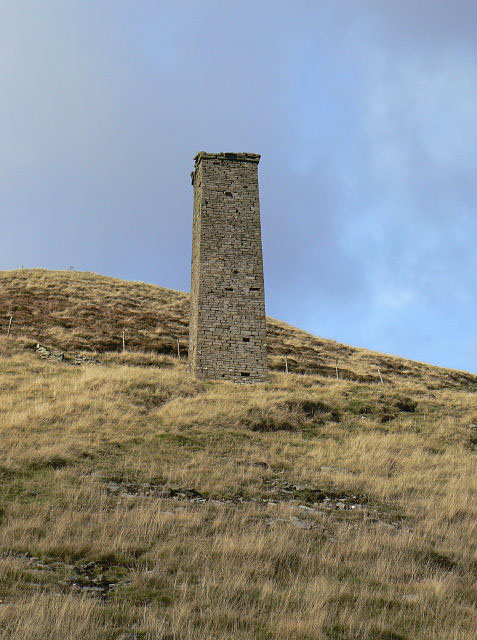 A tall square stone chimney standing alone on a grassy moorland slope under a pale sky.