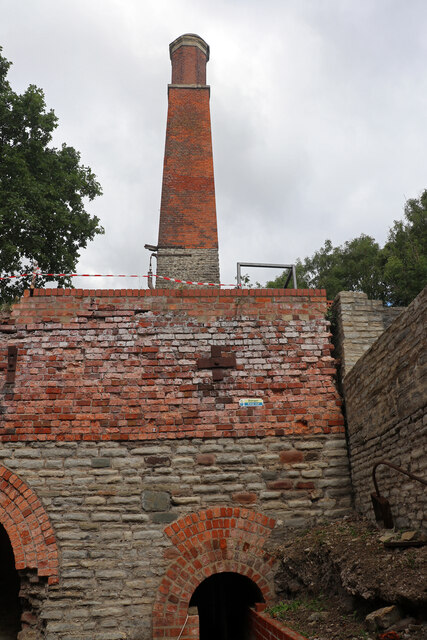 A tall brick chimney rising above a high brick and stone retaining wall with arched openings at its foot, hemmed in by trees.