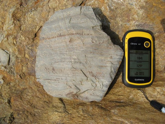 A pale grey slab of rhyolite showing fine banding, laid on orange-stained rock beside a GPS unit for scale.