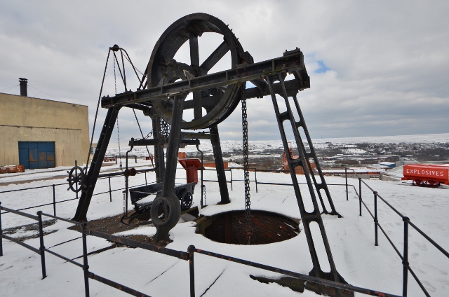 A preserved iron headframe with a large spoked pulley wheel standing over an open shaft in snow, chains hanging from its beam and a small tram beneath it.