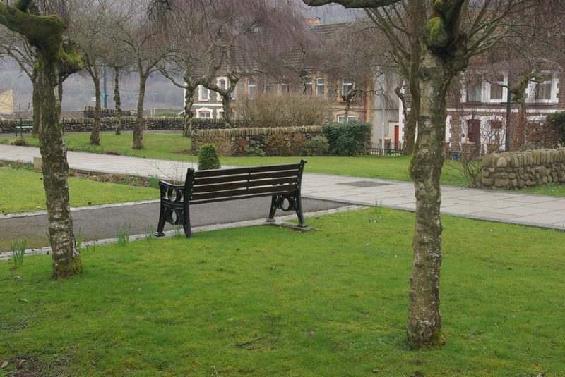 A bench on the grass of a quiet garden among bare trees, with low stone walls and terraced houses behind.