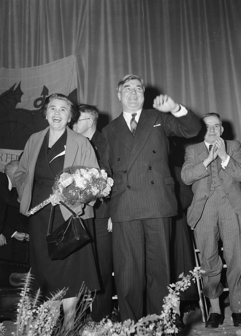 A broad-shouldered man in a pinstriped suit waving from a stage beside a smiling woman holding flowers.