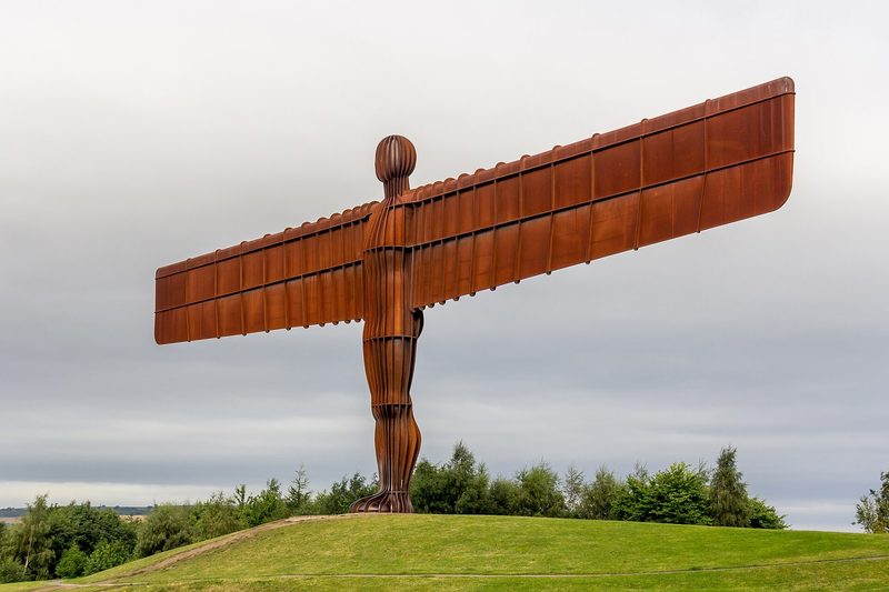 Antony Gormley's Angel of the North, a rust-coloured steel figure with wide horizontal wings, standing on a grassy hilltop against the sky.