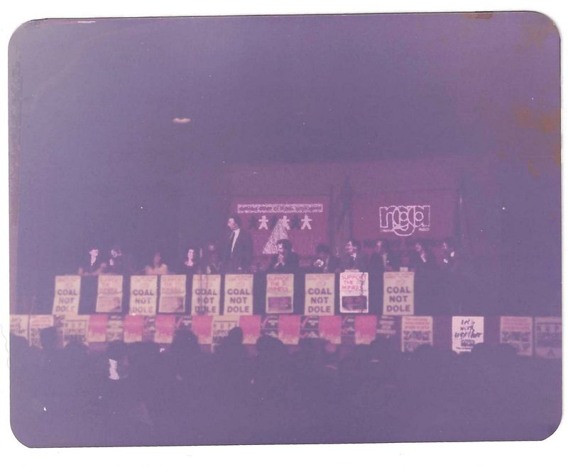 A faded colour photograph of a man speaking from a long platform lined with Coal not Dole placards.