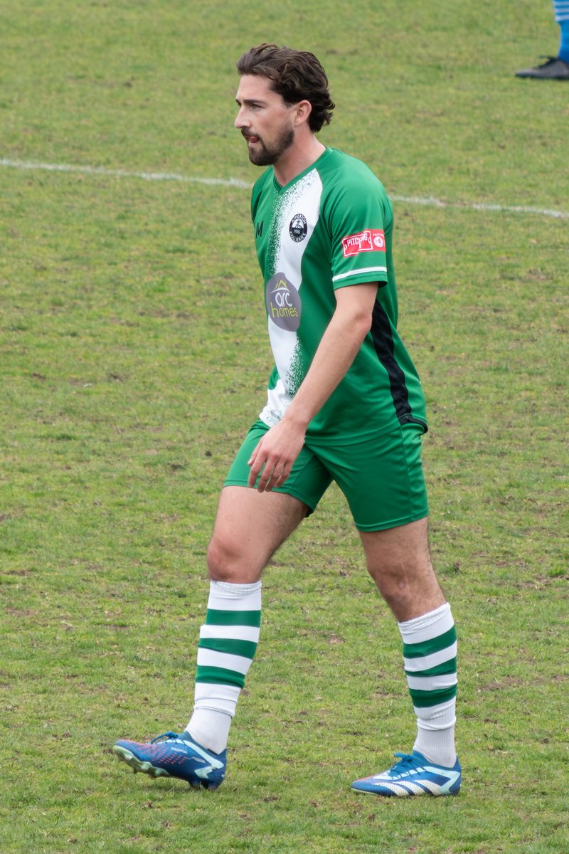A footballer in Atherton Collieries' green and white kit with the club's 1916 badge, walking across the pitch during a 2025 league match
