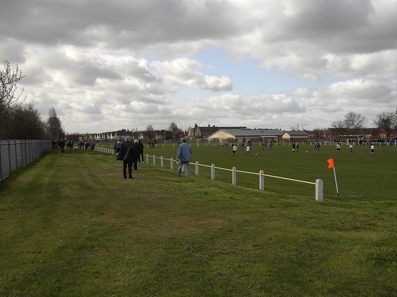 A roped-off football pitch on open ground with spectators standing along the touchline, a corner flag in the foreground and the roofs of the village behind.