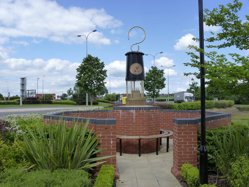 A giant black and brass miner's lamp on a brick pedestal inside a low circular brick wall with a curved bench.