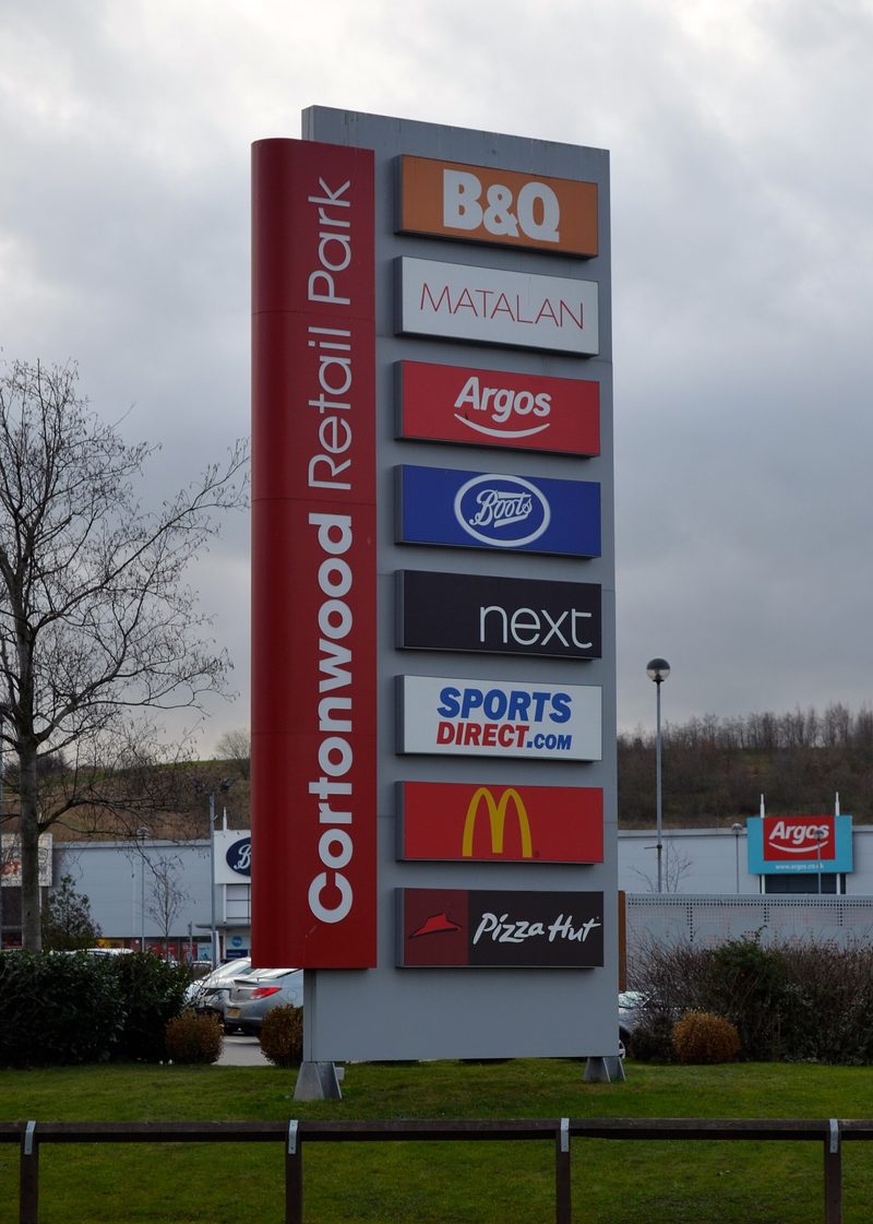 A tall roadside sign for Cortonwood Retail Park listing high-street shops, with store buildings behind.