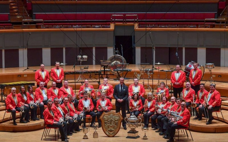 A brass band in red jackets sits on a concert-hall stage with its conductor and a row of trophies in front.