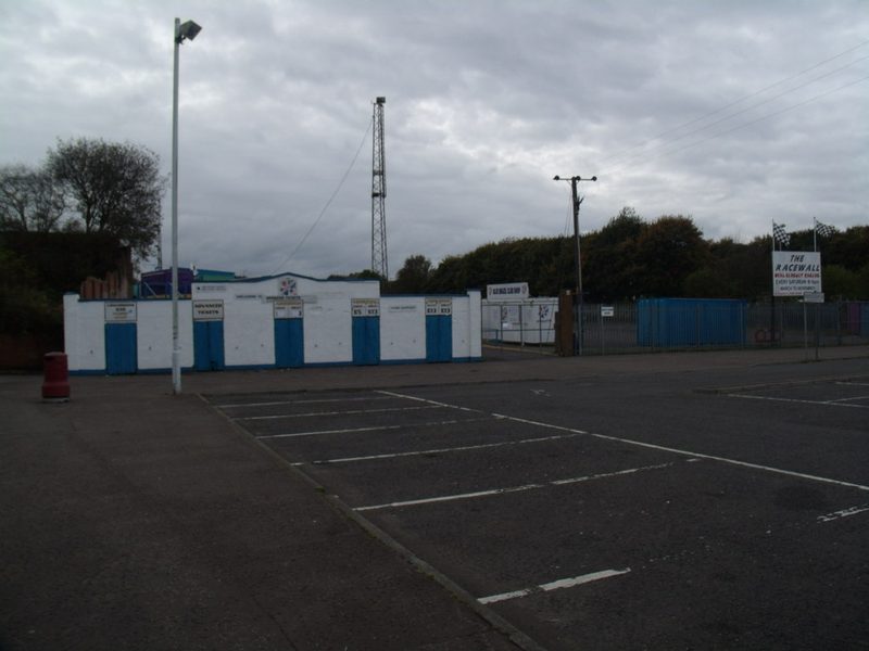 The low white and blue turnstile block of a football ground beside an empty car park, with a floodlight pylon behind.