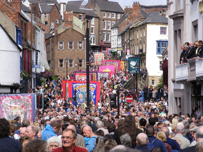A dense crowd fills a Durham street as a line of large, brightly coloured lodge banners is carried downhill between brick buildings.