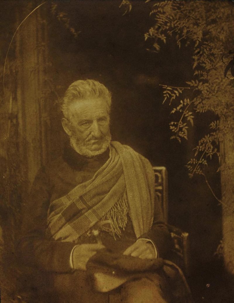A sepia photograph of a white-haired, bearded old man seated in a chair, wrapped in a check shawl, beside a climbing plant.