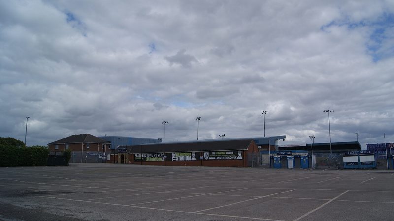 An empty car park before a low brick stand hung with Featherstone Rovers banners, with turnstiles, a ticket office and floodlight pylons under a cloudy sky.