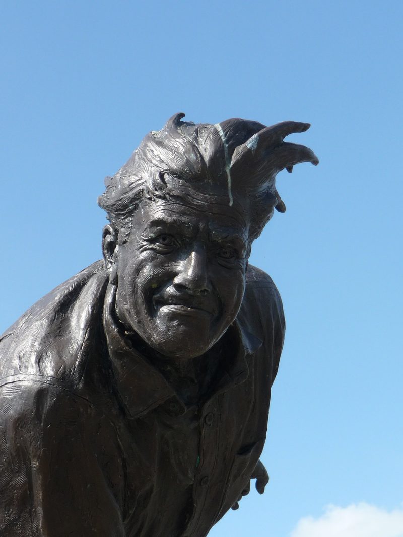 Close view of the head and shoulders of a bronze statue of a smiling bowler with wind-blown hair, against blue sky.