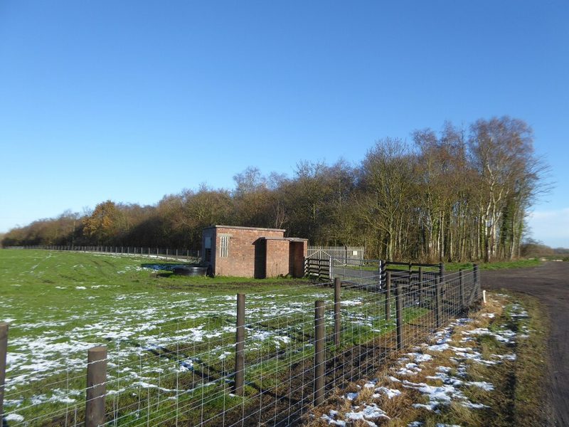 Reclaimed grassland with a small brick building and a fence line in front of a belt of bare trees.
