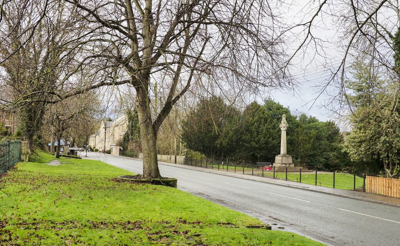 A tall stone wheel-head cross in a railed roadside garden beside a village road lined with bare trees.