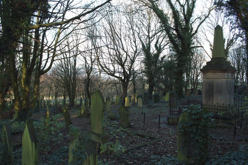 A tall stone obelisk on an inscribed pedestal stands behind iron railings among old headstones in a wooded churchyard.
