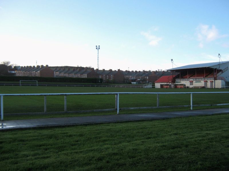 A football ground with a railed-off pitch, a covered main stand with red seats to the right, a floodlight pylon, and rows of terraced houses on the hillside behind.