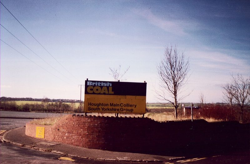A black and yellow British Coal sign reading Houghton Main Colliery, South Yorkshire Group, on a brick wall by a road.