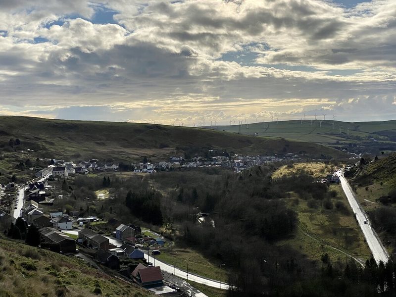 Terraced houses of Gilfach Goch along the floor of a narrow valley, with wind turbines on the ridge beyond.