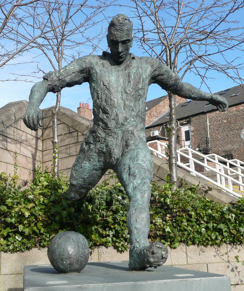 Bronze statue of a footballer in shirt and shorts striking a ball, on a dark stone plinth.