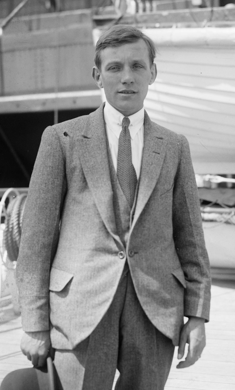 Black-and-white photograph of a slight young man in a light tweed three-piece suit and tie, standing on a ship's deck.