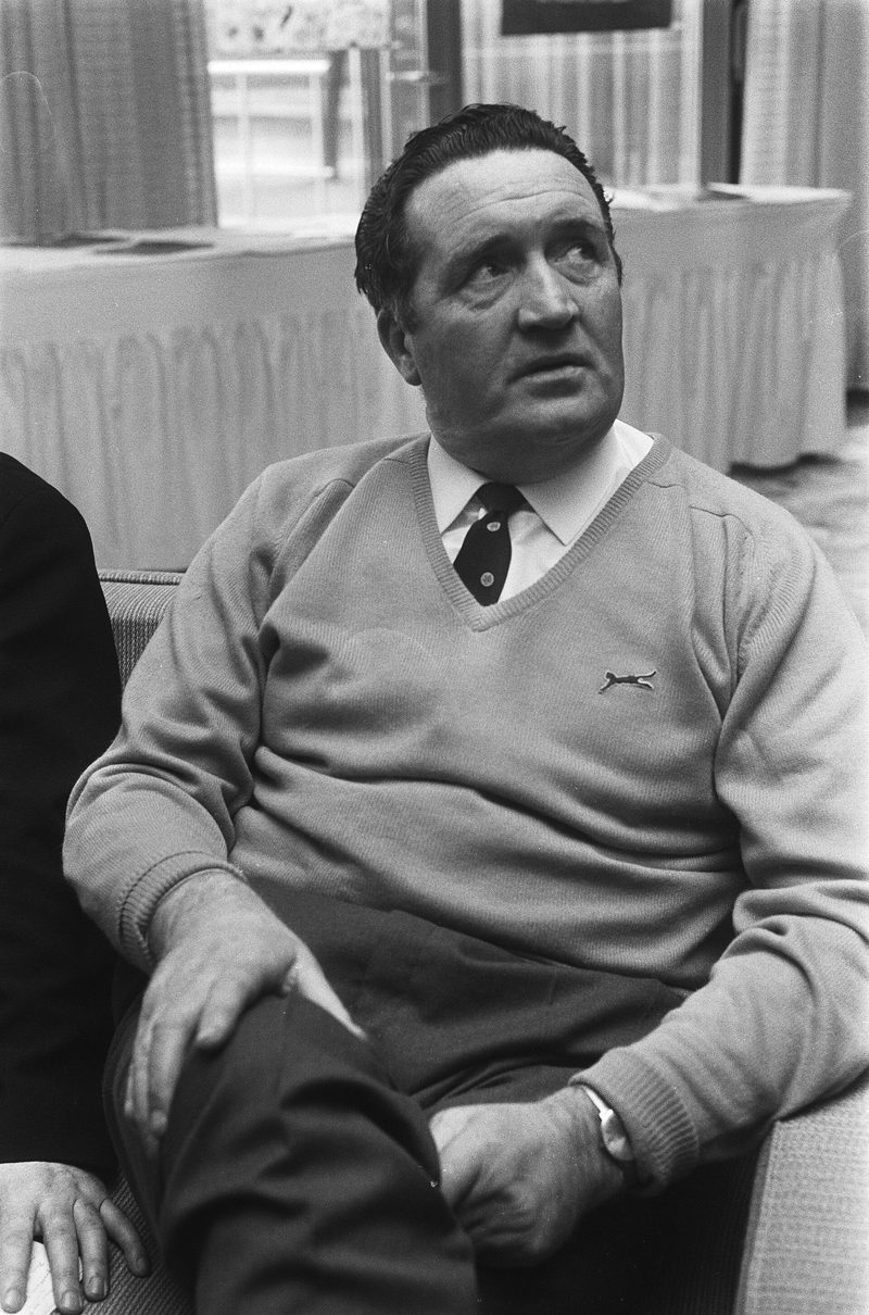 Black and white photograph of Jock Stein seated in an armchair, wearing a light V-neck jumper, shirt and tie, looking up and to one side.
