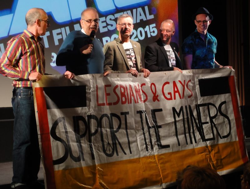 Five men on a cinema stage holding a hand-painted banner reading Lesbians and Gays Support the Miners