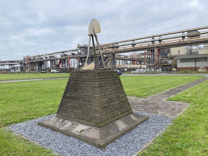 A tapering stone plinth on a lawn carrying crossed miners' shovels in metal, with steelworks pipework and buildings behind it.