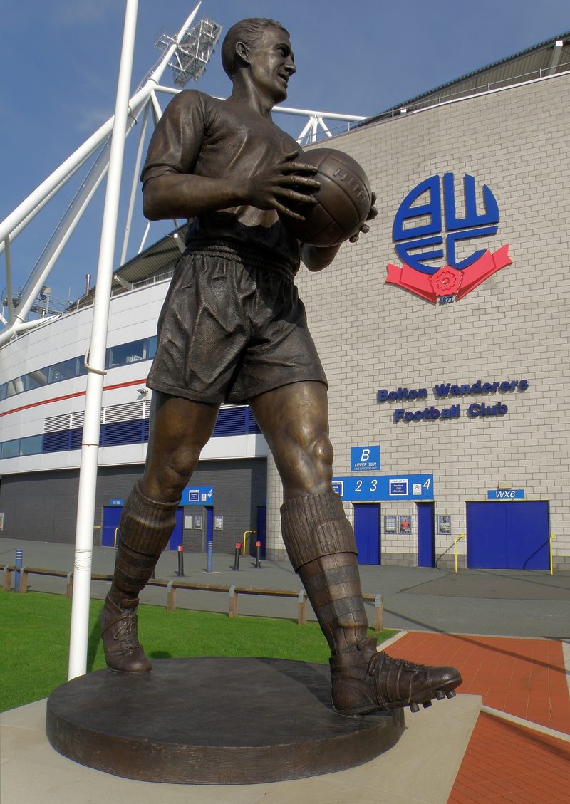 Bronze statue of a footballer in shirt and shorts holding a ball, standing in front of a stadium wall with the Bolton Wanderers crest.