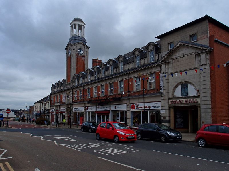 Red-brick Spennymoor Town Hall with its clock tower and a row of shops along the High Street, cars parked in front