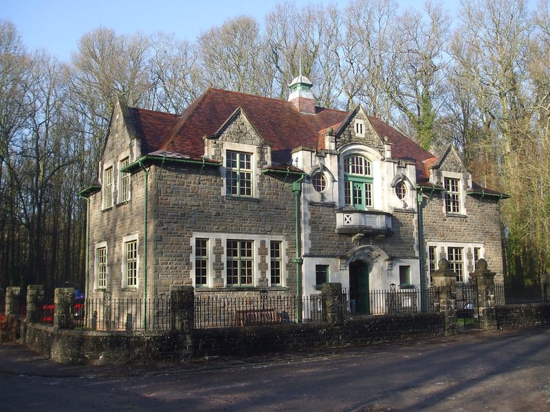 A two-storey stone institute building with gabled dormers, a central bay window over the door and green-painted frames, behind iron railings among bare trees.