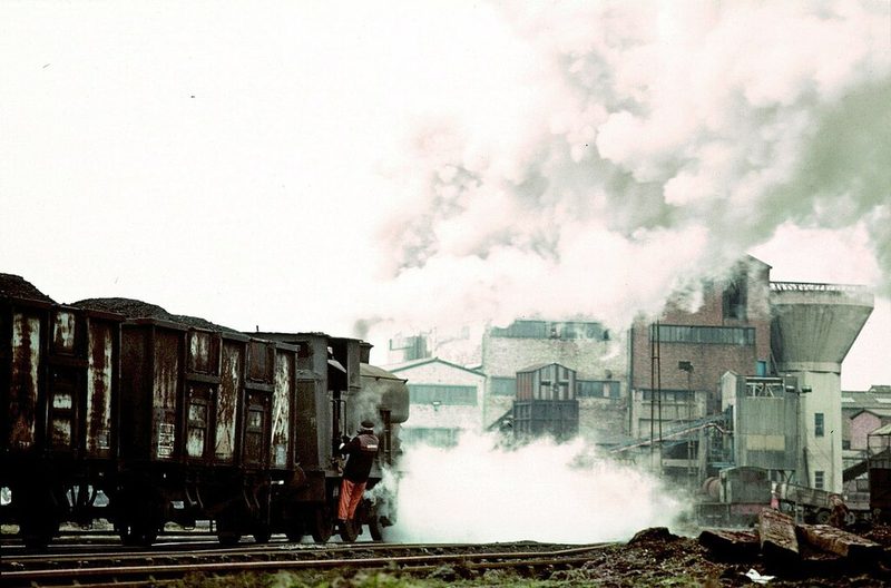 A steam locomotive and loaded coal wagons in clouds of steam in front of colliery buildings and a water tower.