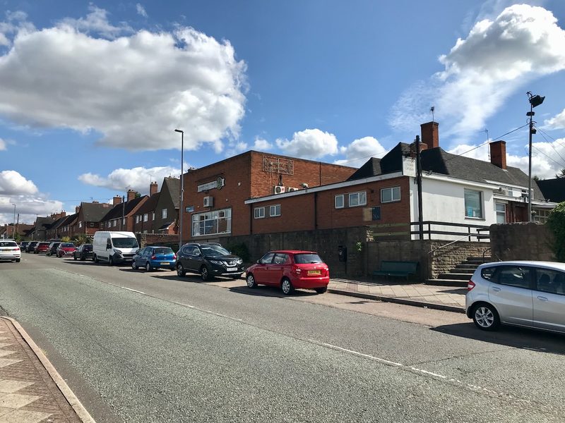 A single-storey brick and rendered social club building beside a village street, with cars parked along the kerb and houses beyond.