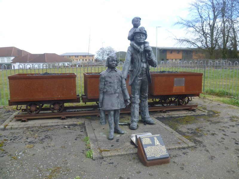 Metal statues of a miner carrying a child on his shoulders and a girl, in front of coal tubs on rails.