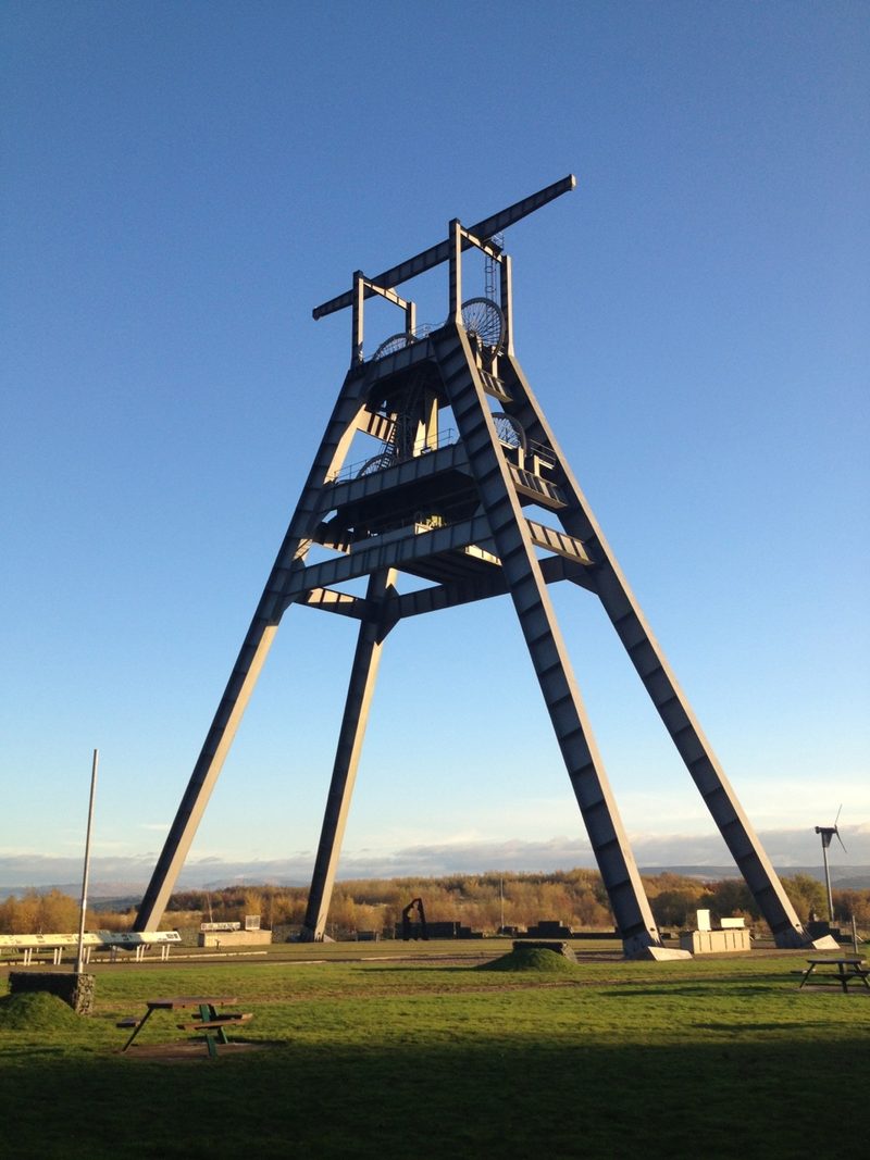 A tall grey steel A-frame headgear with its winding wheels, standing on grass against a clear blue sky.