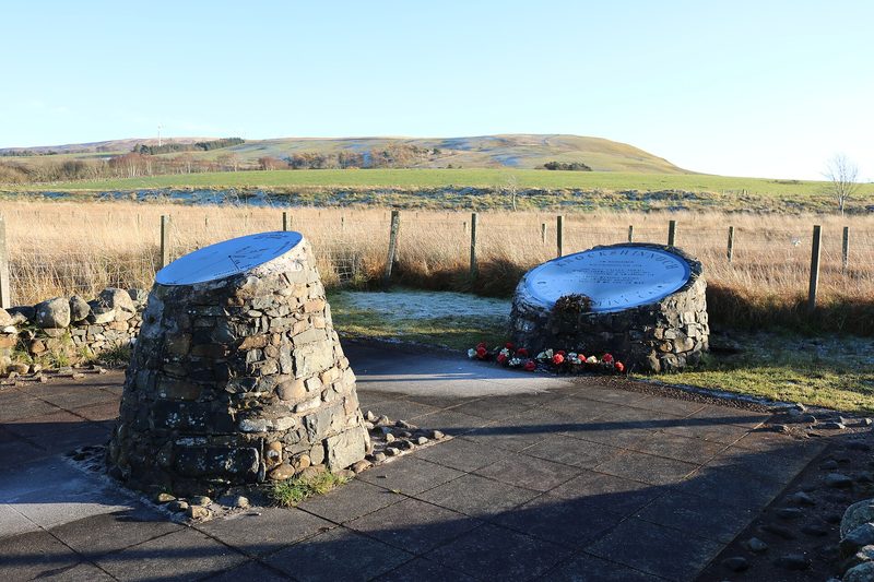 Two stone cairns topped with engraved metal plaques beside a fence, with flowers laid at one and moorland hills behind.