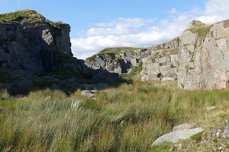 Granite faces of the flooded Foggintor Quarry on Dartmoor rising above rushes and grass under a blue sky.