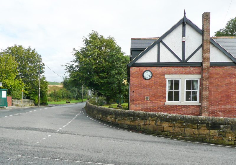 A red-brick building with a mock-Tudor gable and a clock on its wall, standing on a bend of a village road behind a stone wall.